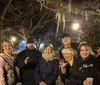 A group of six smiling people are posing for a nighttime photo with some making playful gestures like claws in a park-like setting with trees and street lamps in the background