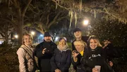 A group of six smiling people are posing for a nighttime photo, with some making playful gestures like claws, in a park-like setting with trees and street lamps in the background.