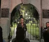 A group of people listens to a woman speaking at the entrance of a historic cemetery at night