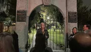 A group of people listens to a woman speaking at the entrance of a historic cemetery at night.