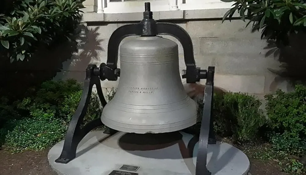 The image depicts a large mounted bell on a concrete base displayed outdoors at night with surrounding foliage