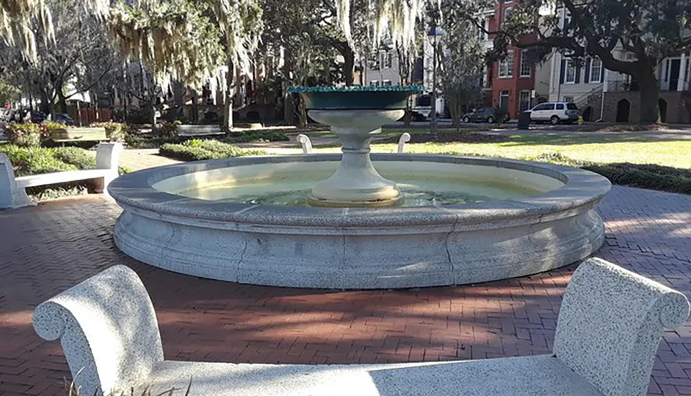 The image shows a large circular fountain with a smaller ornamental basin on top surrounded by benches and trees in an outdoor park setting