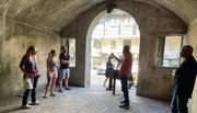 A group of people is attentively listening to a person who appears to be giving a presentation or a tour in an arched, semi-open space with a view of the street outside.