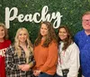Five people are smiling for a photo in front of a green hedge backdrop with the word Peachy in white cursive letters and decorative lights