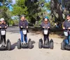 Four people wearing safety helmets are standing on Segways lined up on a path with Spanish moss-laden trees in the background