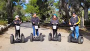 Four people wearing safety helmets are standing on Segways, lined up on a path with Spanish moss-laden trees in the background.