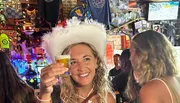 A smiling person wearing a white fluffy hat is toasting with a small glass of beer in a vibrant bar adorned with various decorations and memorabilia.
