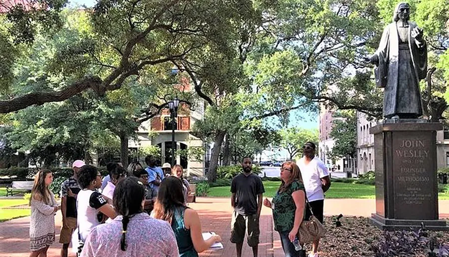 A group of people is gathered around a statue of John Wesley in a park-like setting with lush trees.