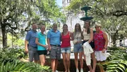 A group of seven people is posing for a photo in a lush park with a fountain in the background, surrounded by large trees draped with Spanish moss.