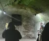 A group of people with a flashlight are exploring a dimly lit arched tunnel with mossy walls which looks like an old historical underground structure