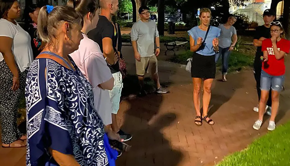 A group of attentive people is gathered around a speaker in an outdoor nighttime setting possibly during a guided tour or event