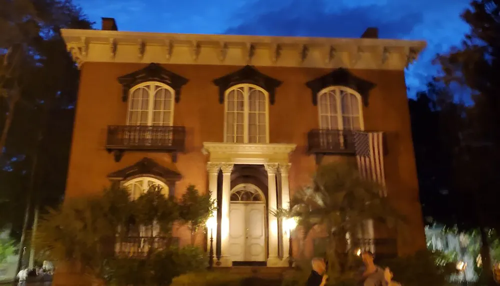 The image depicts a historical two-story brick house at dusk adorned with a lit balcony and an American flag with people gathered out front