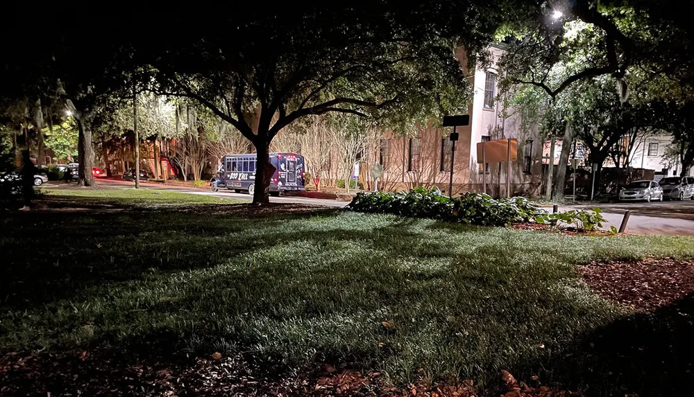 The image captures a nighttime scene of a tranquil urban park area with trees and a trolley bus illuminated by streetlights and a feeling of quiet activity