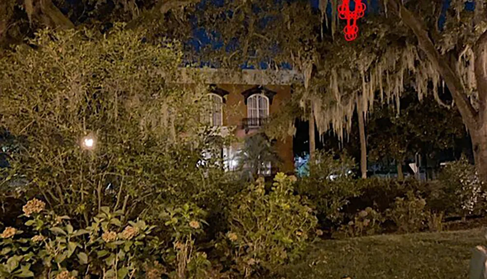 The image shows a spooky-looking house at night partially obscured by trees and Spanish moss with a warm light glowing from one of the windows