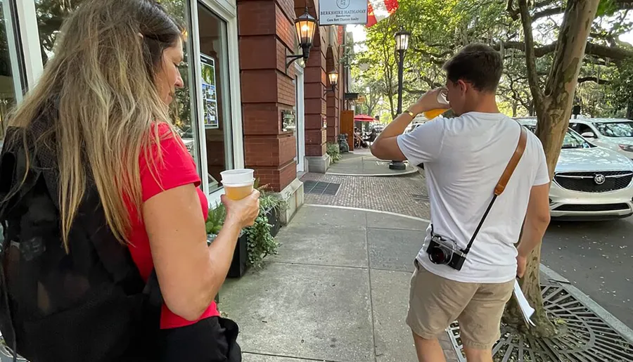 A person holding a coffee cup stands next to another individual, who is holding their phone to their ear, on a city sidewalk with a camera slung over their shoulder.