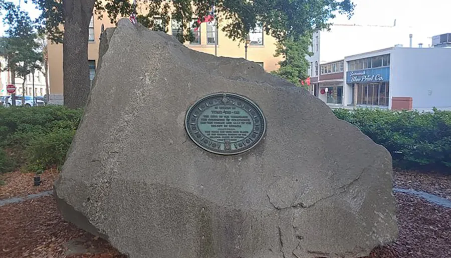 The image shows a large rock with a circular bronze plaque mounted on it, set within an urban park or garden area.