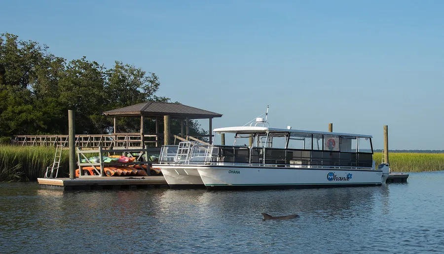 A pontoon boat named Ohana is docked at a wooden pier on a calm body of water with a gazebo and kayaks nearby, under a clear blue sky.