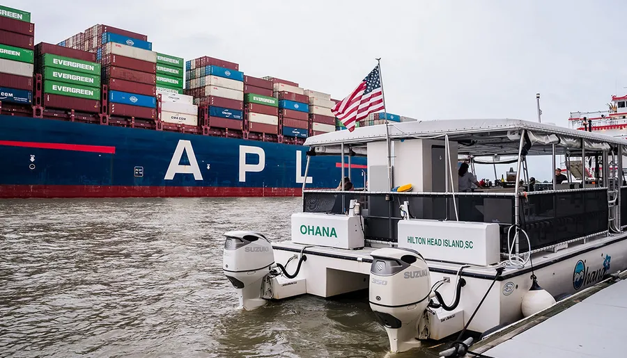 A pontoon boat named OHANA flies the American flag as it travels near a large cargo ship loaded with containers.