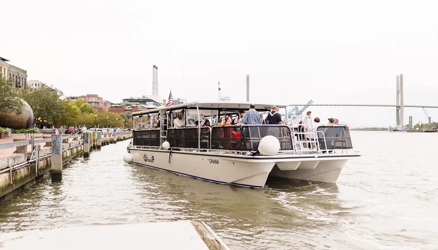 A group of people is enjoying a ride on a riverboat named Ohana that is docked along a city waterfront.