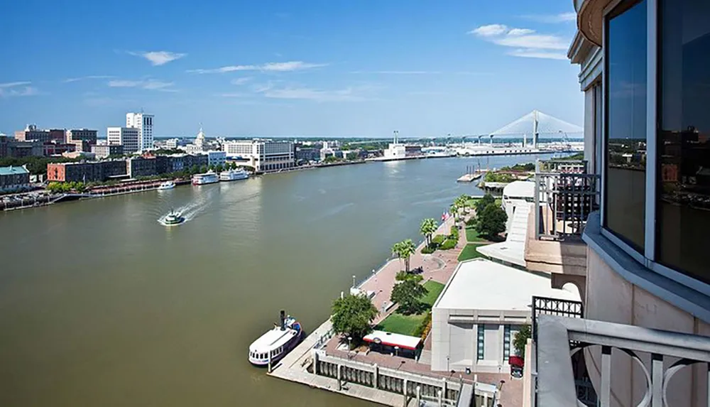 This is a riverside urban landscape featuring a wide river with boats a waterfront promenade with trees and a variety of buildings including a distant bridge in the background