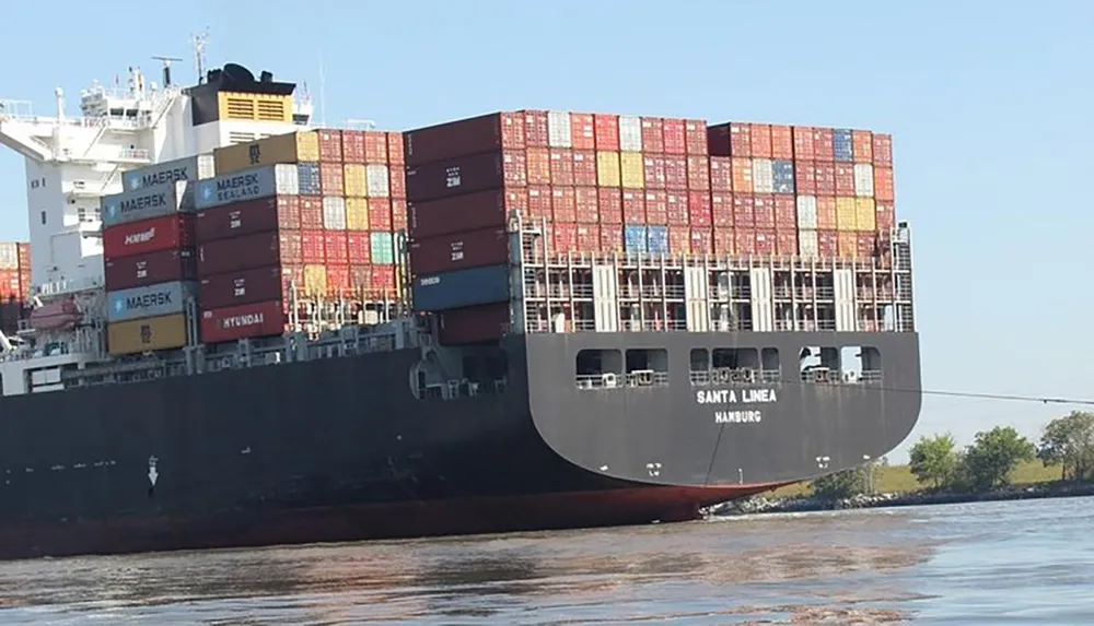 The image shows a large cargo ship heavily loaded with stacked shipping containers sailing on the water