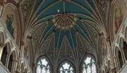The image shows the intricate, vaulted ceiling and stained glass windows of a Gothic-style church interior, adorned with colorful patterns and religious iconography.
