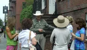 A person wearing a wide-brimmed hat and a uniform is speaking to a group of attentive women on a city street.