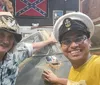 Two smiling men one wearing a military-style hat with badges and another in a USN United States Navy cap pose for a selfie in front of what appears to be a part of a military aircraft in a room adorned with military memorabilia