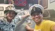 Two smiling men, one wearing a military-style hat with badges and another in a USN (United States Navy) cap, pose for a selfie in front of what appears to be a part of a military aircraft, in a room adorned with military memorabilia.