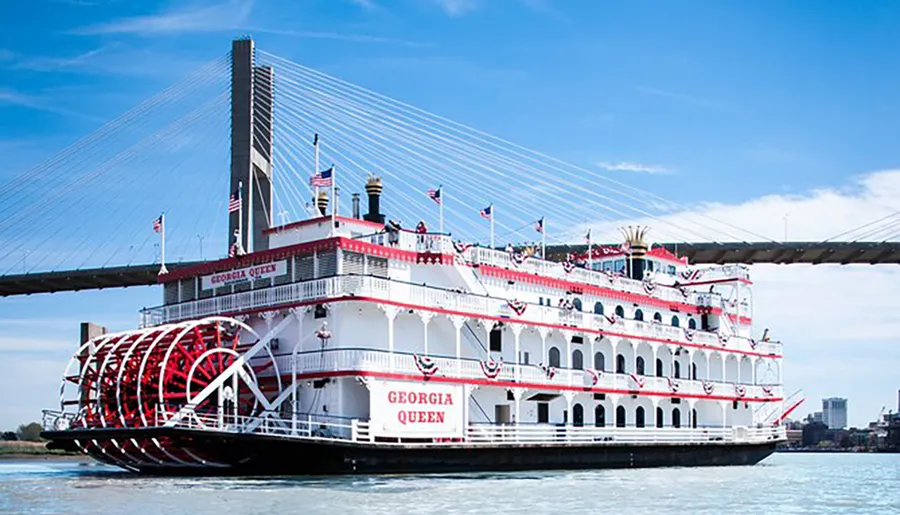 The image features the Georgia Queen, a large paddle wheel riverboat, festooned with American flags, cruising on a river near a modern cable-stayed bridge on a sunny day.