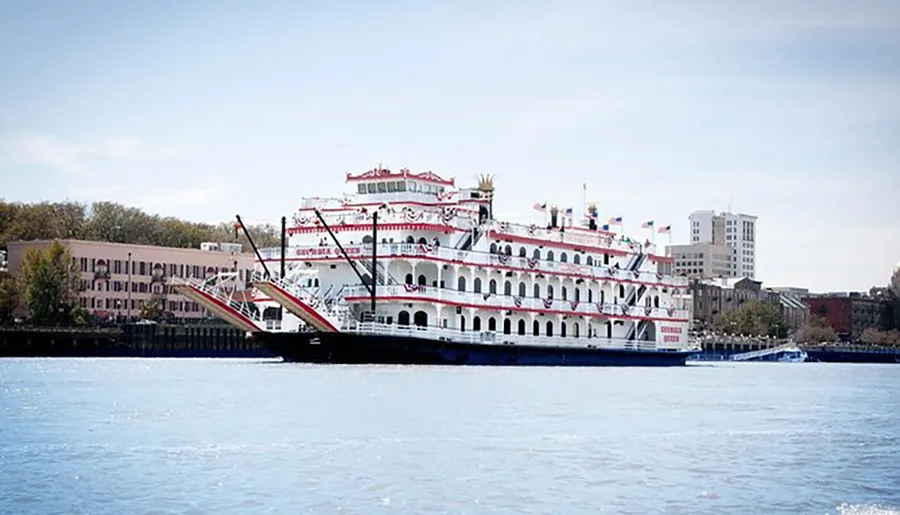 A traditional paddle steamer with multiple decks and red accents is docked on a river along an urban waterfront.