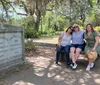 Three individuals are posing for a photo while sitting on a bench in a park-like environment with draped Spanish moss and a historic gravestone to the left