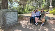 Three individuals are posing for a photo while sitting on a bench in a park-like environment with draped Spanish moss and a historic gravestone to the left.