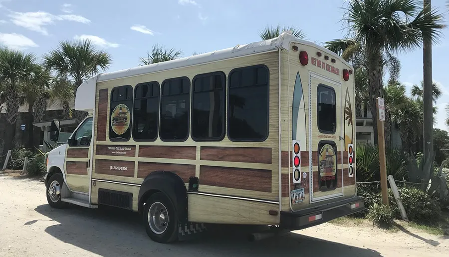 A trolley bus with wood paneling design is parked near palm trees, displaying advertising and contact information for what appears to be a tour or shuttle service.