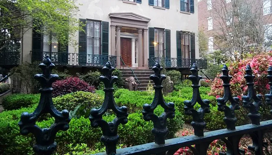 The image shows a classic townhouse with a balcony and shutters, viewed through an ornate black iron fence surrounded by lush greenery.