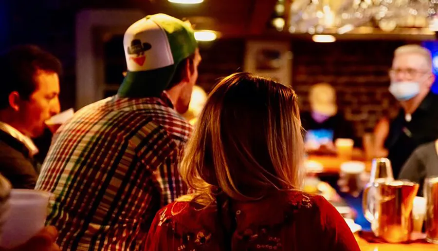 People are socializing at a dimly lit bar with some wearing masks, suggesting precautions during a time of health concerns.