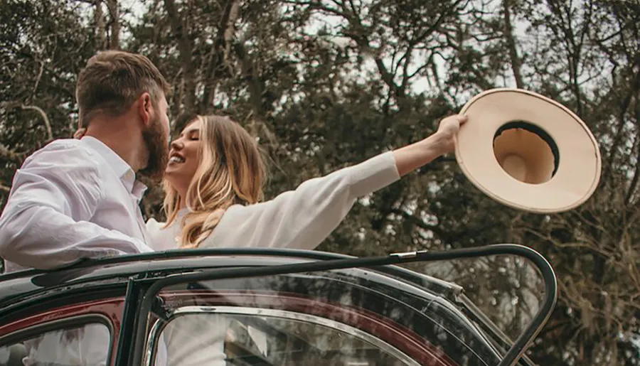 A joyous couple is sharing an intimate moment, with the woman raising her hat in a celebratory gesture, while standing through the sunroof of a car amid a backdrop of trees.