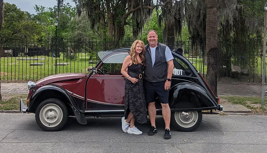 A man and a woman are smiling and posing in front of a classic two-toned car in a park-like setting with trees and Spanish moss in the background.