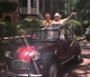 Two people are joyfully poking out of the sunroof of a classic red car parked on a picturesque tree-lined street with brick pavement