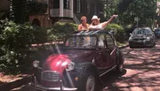 Two people are joyfully poking out of the sunroof of a classic red car parked on a picturesque tree-lined street with brick pavement.