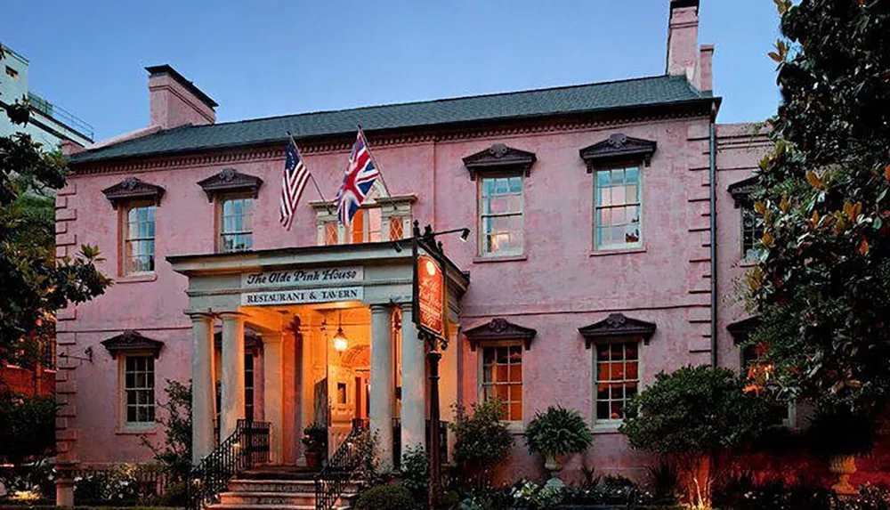 This image shows the facade of The Olde Pink House an elegant pink-colored historical building housing a restaurant and tavern flying both the American and British flags at dusk