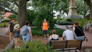 A group of people is attentively listening to a tour guide who is standing in front of them in an urban park setting.