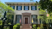 The image shows an elegant, traditional two-story building with green window shutters, a central entrance flanked by flags, and a fenced, leafy front yard.