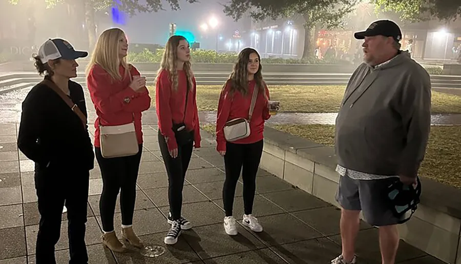 Four people, three wearing red tops, are attentively listening to a man as they stand on a city sidewalk against a foggy background at night.