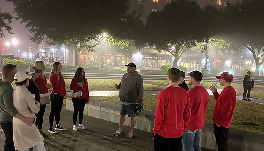 A group of people, some wearing matching red tops, are listening to a person who appears to be addressing them in a foggy, outdoor urban setting at nighttime.