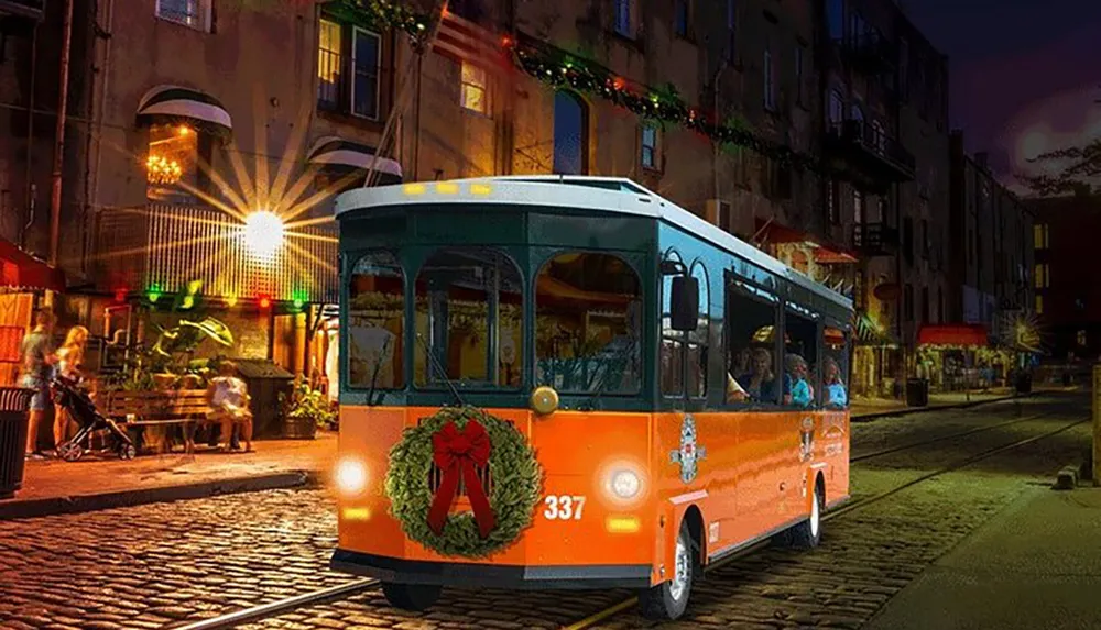 An orange vintage-style trolley adorned with a Christmas wreath travels down a festive cobblestone street at night