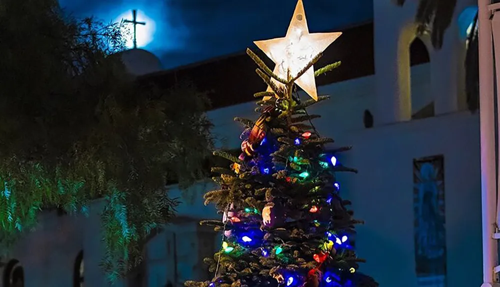 A Christmas tree adorned with colorful lights stands before a church with a glowing cross in the evening