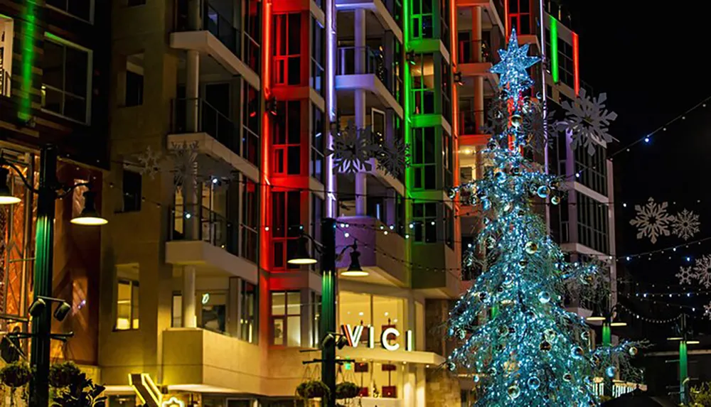 The image shows a vibrant night scene of a festive outdoor area with an illuminated Christmas tree and colorful building balconies suggesting a holiday celebration in an urban setting