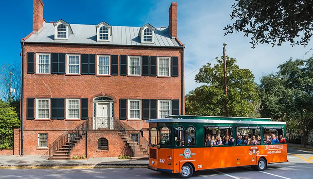 A trolley full of passengers is driving by a large red-brick colonial-style building on a sunny day