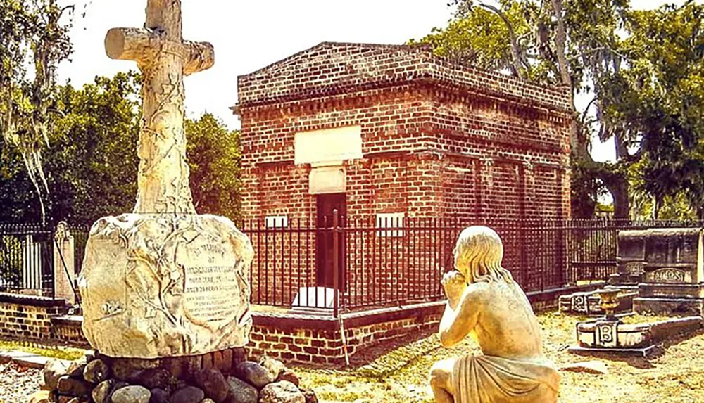 The image depicts a tranquil cemetery setting with a large cross monument and a statue of a person kneeling in prayer before a historical brick mausoleum surrounded by mature trees and grave markers